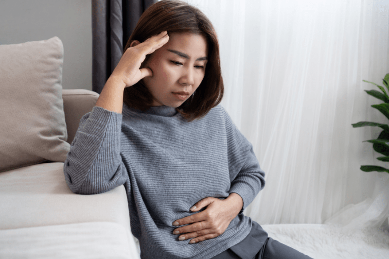 Woman sitting with discomfort, holding her abdomen and showing signs of digestive upset or stomach pain.